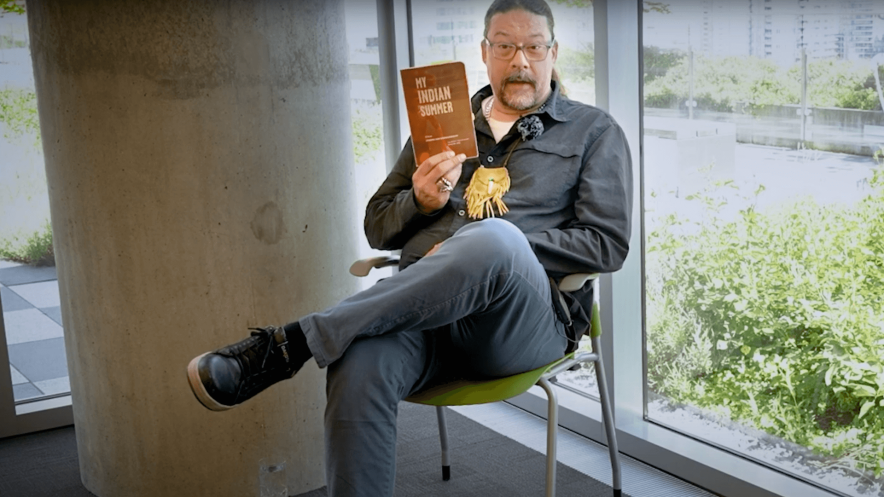 Author Joseph Kakwinokanasum sits cross-legged on a green chair by a large window, holding up a copy of his book My Indian Summer. He is wearing a dark button-up shirt, light blue jeans, black boots, and a fringed necklace, with a concrete pillar and lush greenery visible in the background.